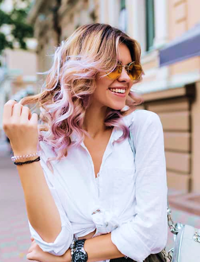 A stylish woman with vibrant hair and a bright smile, possibly enhanced by cosmetic dentistry, poses outdoors in this photo.